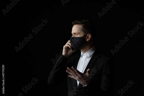 Excited and stressed businessman wearing a face mask and a suit speaking on smartphone. Isolated against a black background.