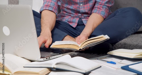 Unrecognizable young man studying online or doing homework using laptop and books sitting on floor at home
