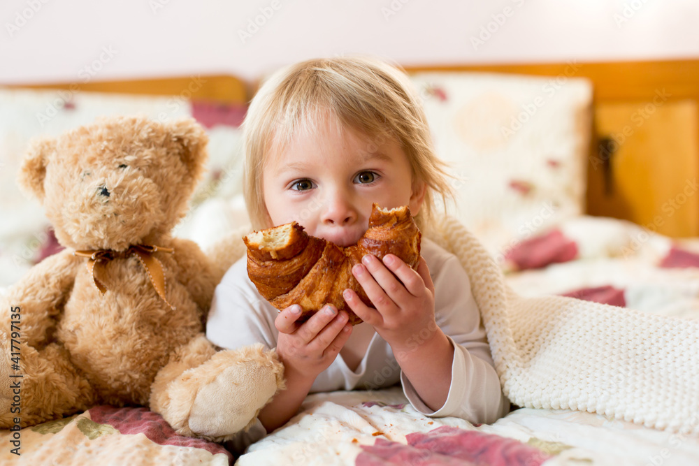 Cute blonde toddler child,reading book and eating croissant in bed, laughing Cute blonde toddler child,reading book and eating croissant in bed, laughing