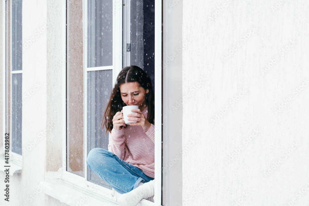 Beautiful brunette woman sitting on a window sill  at home and drinking hot drink, cozy winter vacations portrait