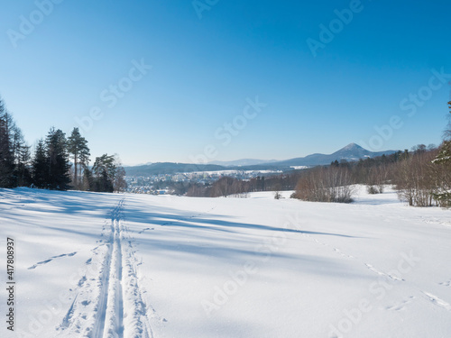 Winter landscape with view of village town Cvikov and ski run trail on snow-covered fields and snowy frozen forest and trees on sunny day, blue sky background © Kristyna