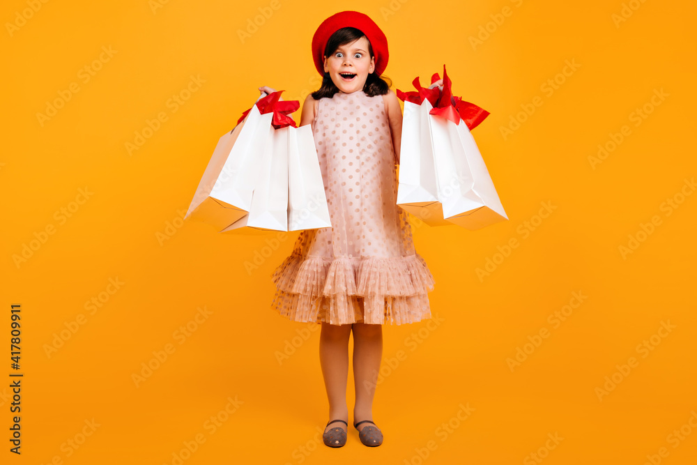 Fototapeta premium Surprised little girl in french beret posing after shopping. Studio shot of amazed kid with paper bags.