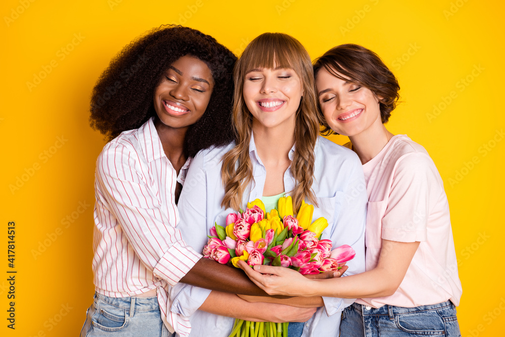 Portrait of three attractive trendy dreamy cheerful girls bonding embracing holding tulips isolated over bright yellow color background Portrait of three attractive trendy dreamy cheerful girls bonding embracing holding tulips isolated over bright yellow color background