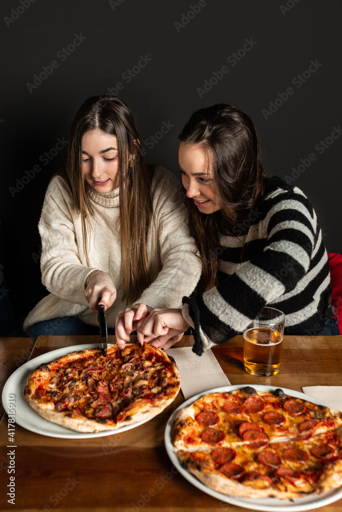 two girls at a restaurant table cutting a pizza with a pizza cutter. two girls at a restaurant table cutting a pizza with a pizza cutter.