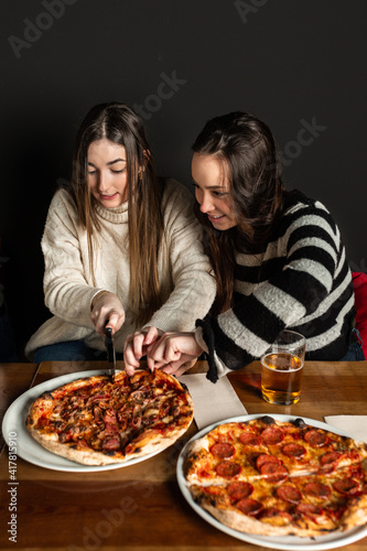 two girls at a restaurant table cutting a pizza with a pizza cutter.