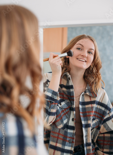 Cute Caucasian woman using a makeup brush and smiling in front of the mirror over home background. Beauty, make up, cosmetics, morning and people concept