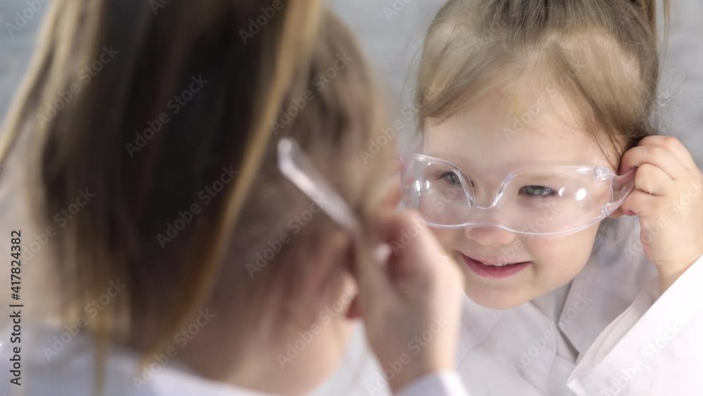 A girl in a white coat is playing a doctor, trying on glasses in front of a mirror. Emotions of children. A girl in a white coat is playing a doctor, trying on glasses in front of a mirror. Emotions of children.