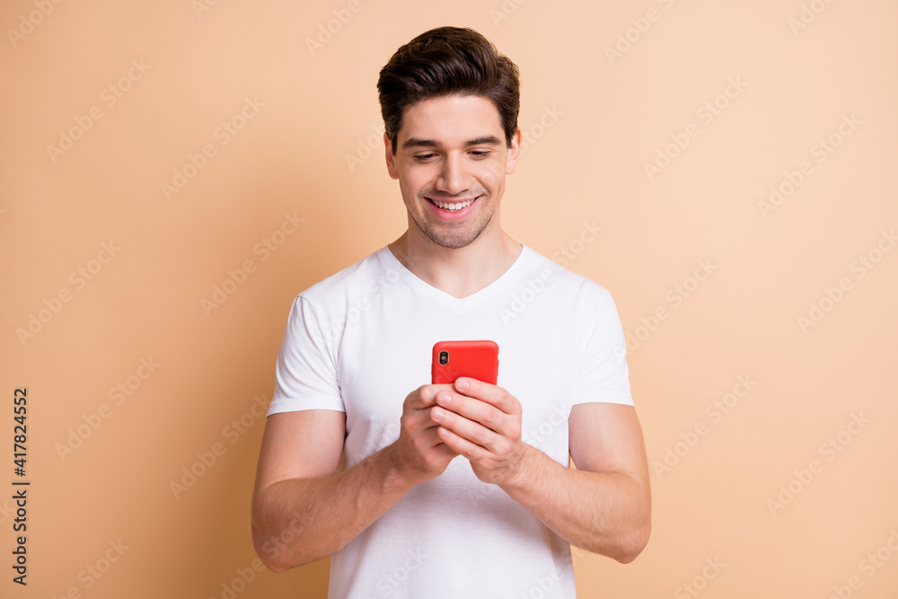 Photo of handsome young person hold phone look screen toothy smile isolated on beige color background Photo of handsome young person hold phone look screen toothy smile isolated on beige color background