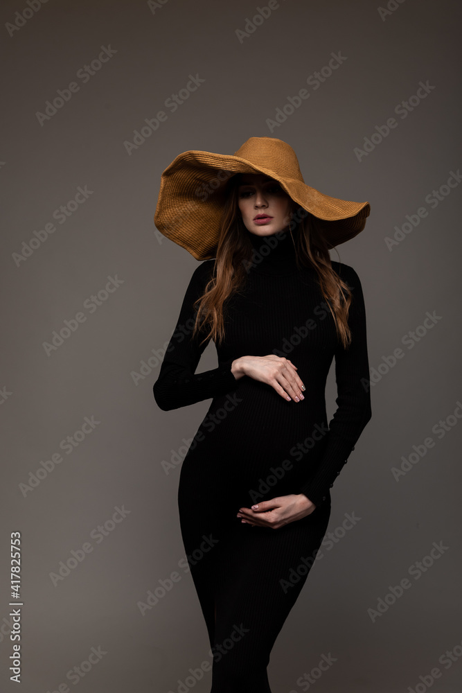 Pregnant girl in black tight dress. In a big hat. Hands on belly. Looking down. Grey background. Pregnant girl in black tight dress. In a big hat. Hands on belly. Looking down. Grey background.