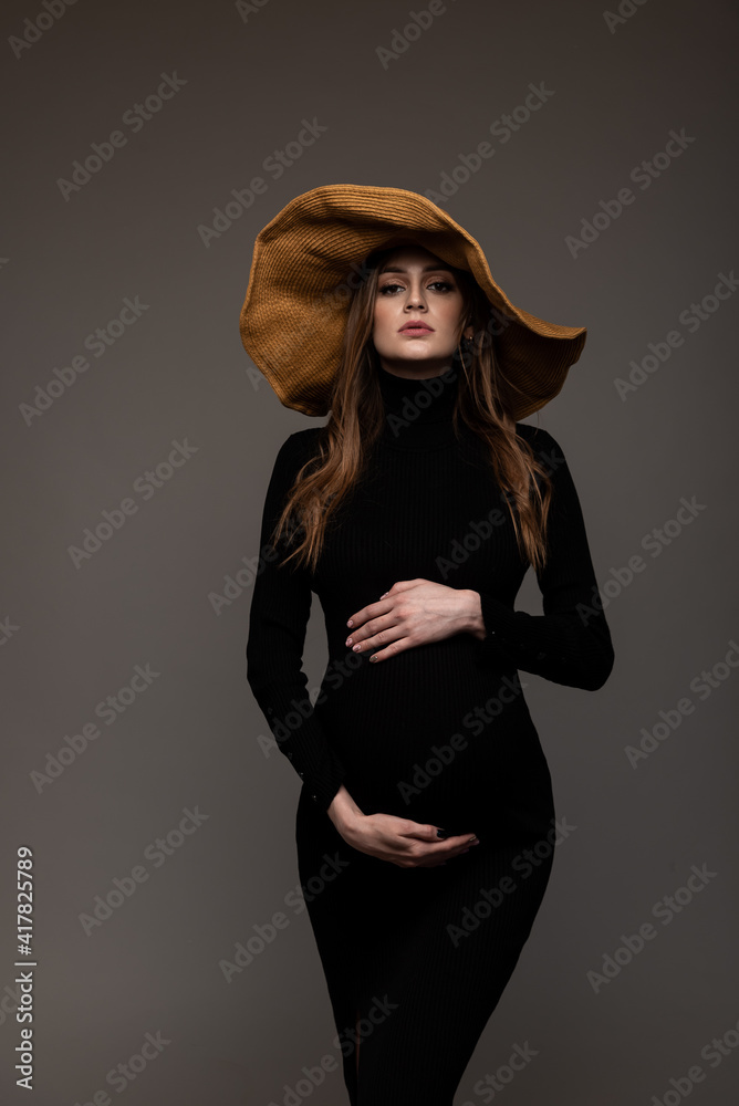 Pregnant girl in black tight dress. In a big hat. Hands on belly. Grey background. Pregnant girl in black tight dress. In a big hat. Hands on belly. Grey background.