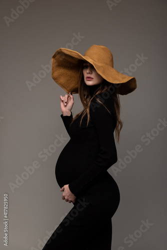 Pregnant girl in black tight dress. In a big hat. Profile. Hand holding hat. Grey background.