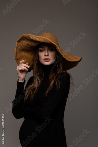 Portrait pregnant girl in black tight dress. In a big brown hat. Hand to face. Grey background.