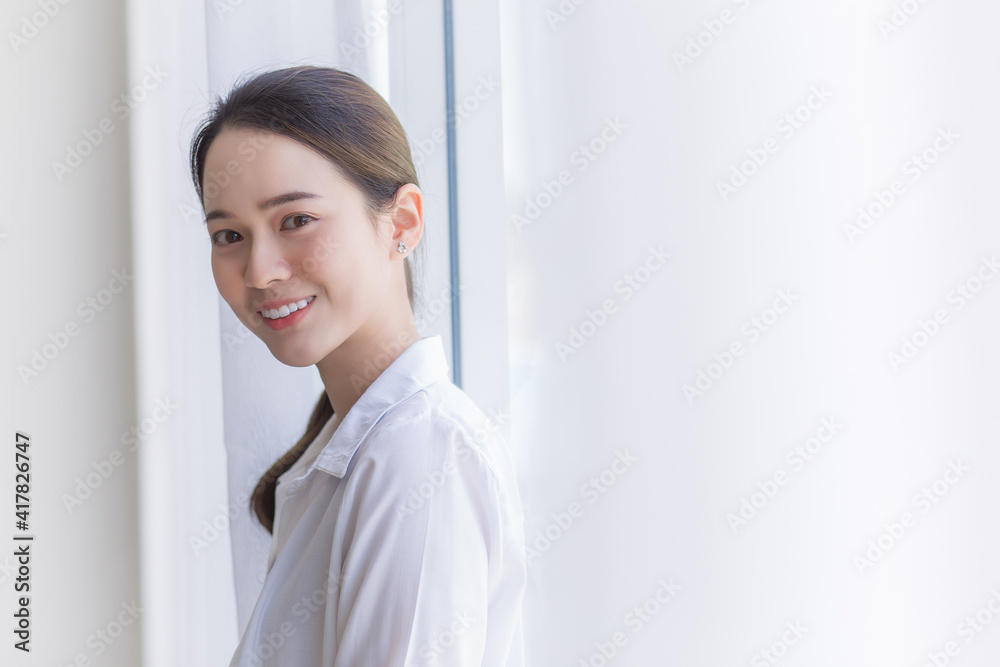 Asian beautiful woman in white shirt is smiling and standing near window with white curtain. Asian beautiful woman in white shirt is smiling and standing near window with white curtain.