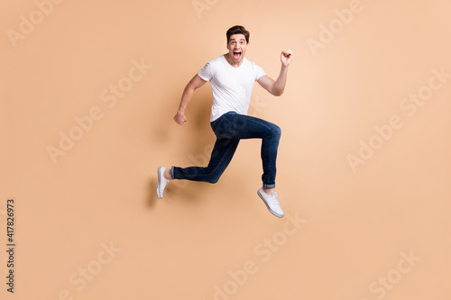 Full size profile photo of hooray brunet man jump run yell wear t-shirt jeans sneakers isolated on beige background