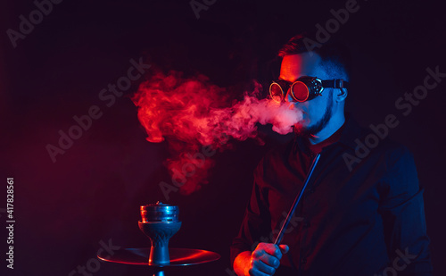man in futuristic glasses smokes a hookah and blows a cloud of smoke in a shisha bar