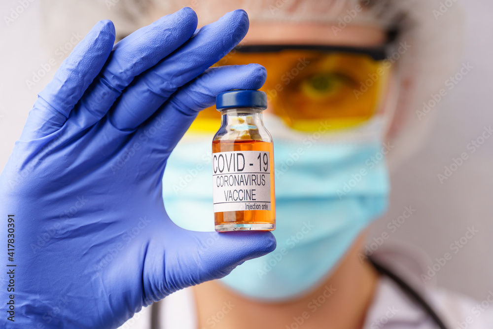 Female doctor in a protective mask and glasses holds a vaccine bottle in her hand,