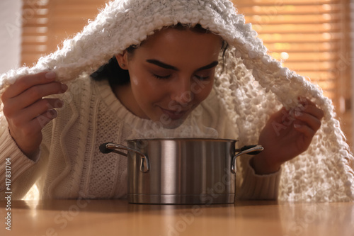 Woman with plaid doing inhalation above saucepot at table indoors photo