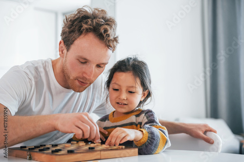 Single father with his daughter is at home together at daytime. Playing checkers