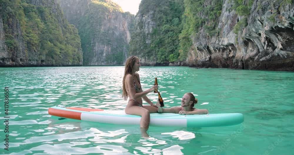 Happy young romantic caucasian couple male and female sitting on stand up paddle board enjoying summer vacation having fun celebrating summertime holiday with smiling face drinking beer and kissing.
