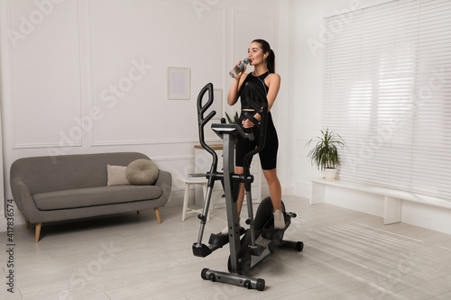 Happy young woman drinking water during training on elliptical machine at home