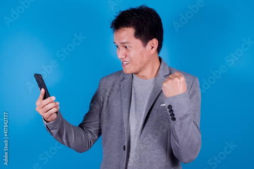 Asian man at cellphone and looking at camera with amazement on blue background. Man shows winning gesture by raising his hand.