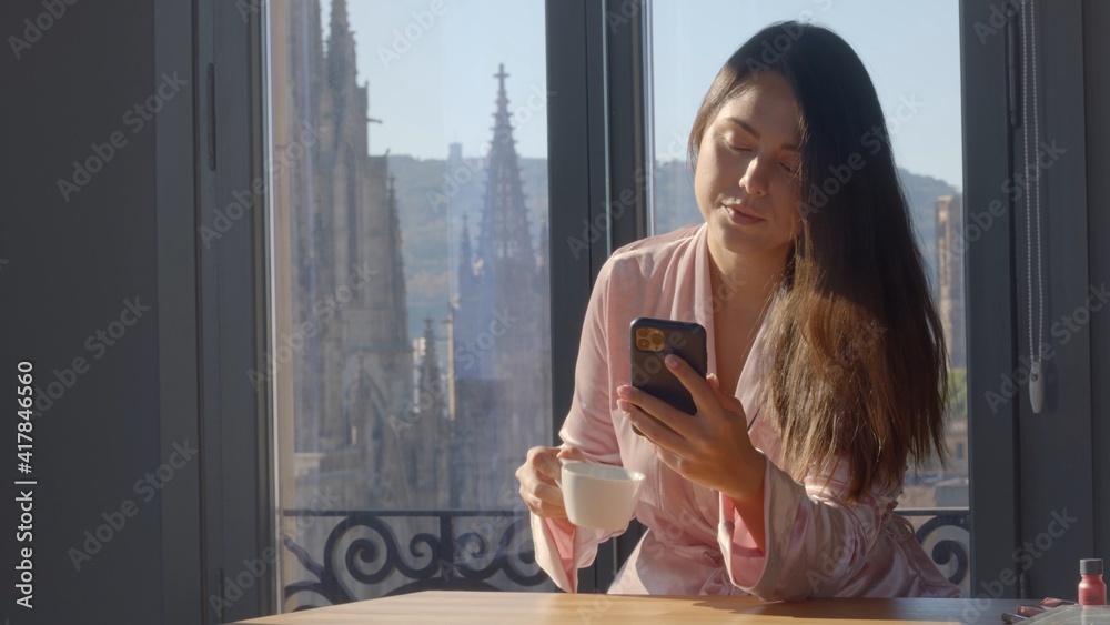 Beautiful caucasian girl with brown straight hair at home with Barcelona view. Velvet light bathrobe. Enjoying morning coffee. Watching her phone, smiling. Sunny day. High-quality medium shot portrait