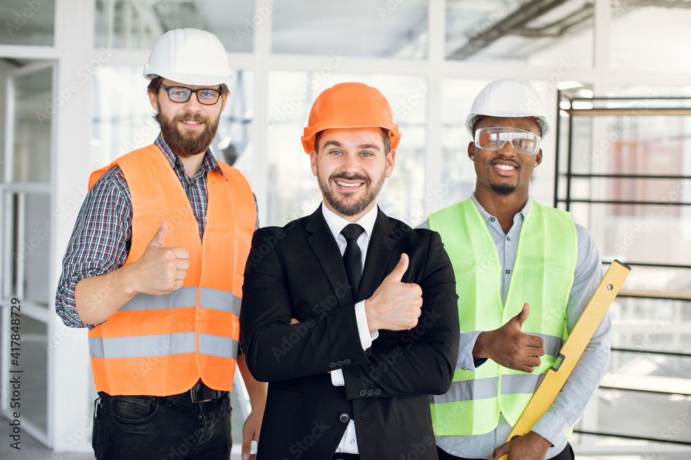Multicultural partners smiling and showing thumbs up. Three men wearing protective helmets and glasses. Success of construction business. Multicultural partners smiling and showing thumbs up. Three men wearing protective helmets and glasses. Success of construction business.