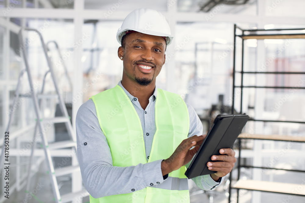 Smiling afro-american engineer in construction hardhat working on digital tablet. Positive man in uniform using modern gadget at work. Business person. Smiling afro-american engineer in construction hardhat working on digital tablet. Positive man in uniform using modern gadget at work. Business person.