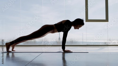 Slow motion Tracking shot of a female slim yoga instructor on the floor of a big pilates studio with big windows and views of blue sky and sea, stretching with fitness routine, sun salutation.
