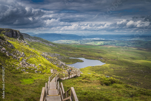 People hiking on steep stairs of wooden boardwalk in Cuilcagh Mountain Park. View on lake and valley below with dramatic sky, Northern Ireland photo