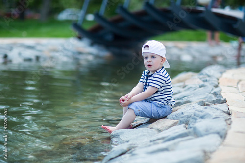 Baby in a white cap sitting by  pond touches cold water with his leg and admires