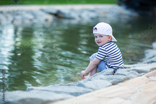 Fototapeta Naklejka Na Ścianę i Meble -  Baby in a white cap sitting by the pond touches cold water with his leg and admires