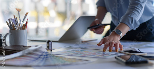 Graphic designer holds a smartphone with color charts and office equipment on work desk.