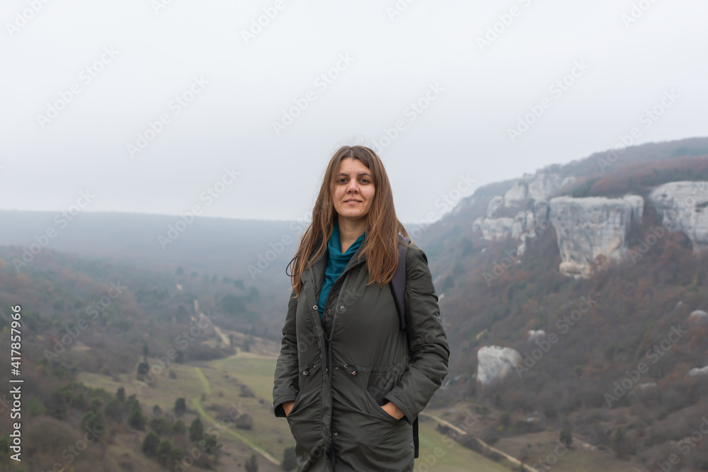 A young dark-haired woman on the background of an autumn landscape. Cloudy gray foggy view from the mountain. A happy smiling girl in a green coat. Natural background, concept of hiking, ecotourism