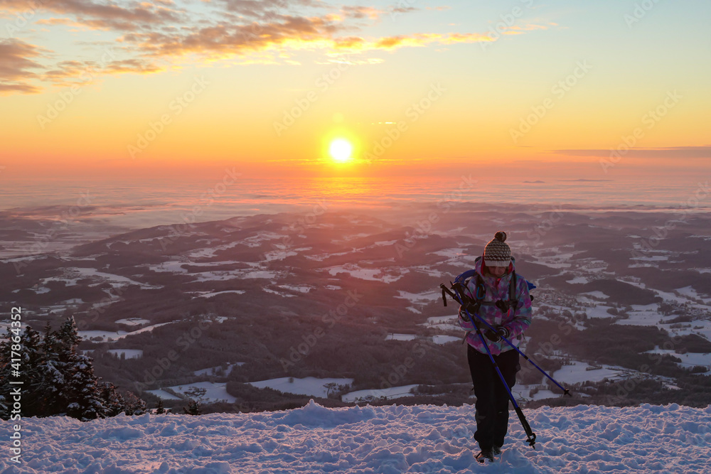 Woman hiking to the top of snow covered peak of Schoeckl in Austrian Alps and admiring a sun rising above the horizon. The sky is bursting orange, trees sprinkled with snow. Winter landscape. Daybreak