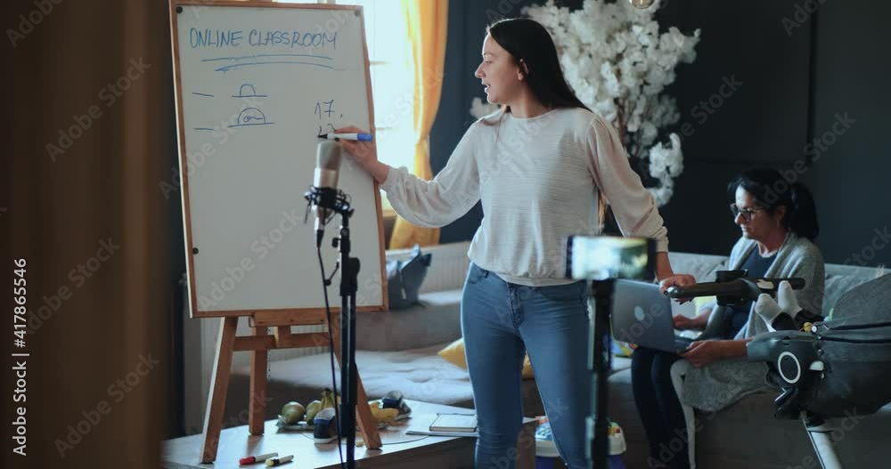 Capable young mother looks after her child and works from home remotely using a live stream, a school blackboard in the living room. Granny woman in the background typing on laptop keyboard, working