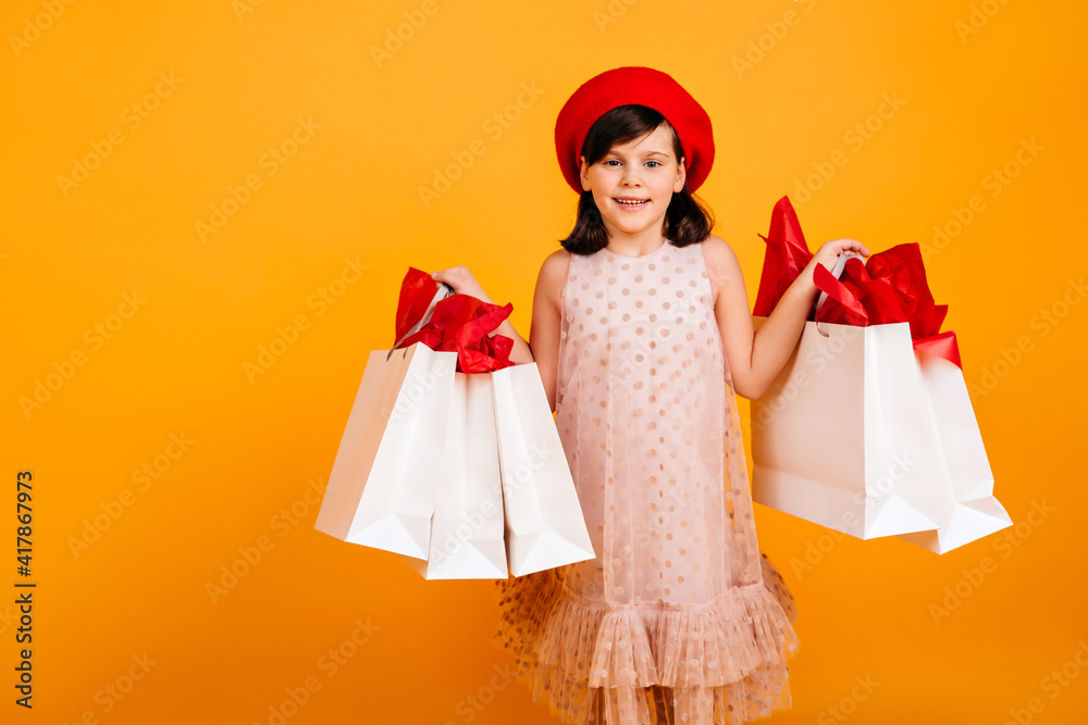 Joyful girl in elegant dress smiling on yellow background. Cute child holding store bags and looking at camera.