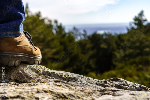 Detail of a man's shoe perched on a rock in front of the green mountain landscape and the blue sky.