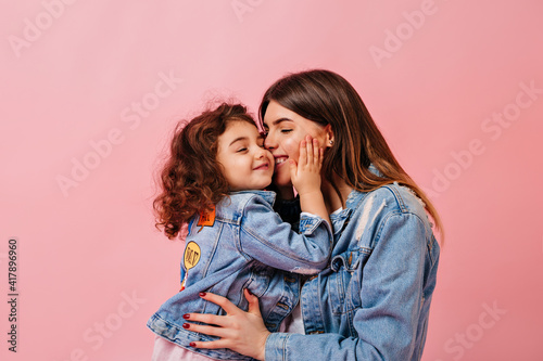 Relaxed preteen girl embracing with mother. Winsome young mom kissing daughter on pink background.