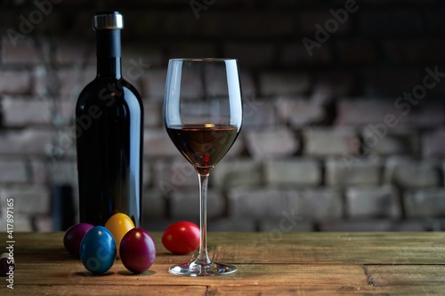 Easter decorations, colorful eggs, a glass of red wine with a bottle on table at home dark warm living room, brick wall in background. Copy space for text.