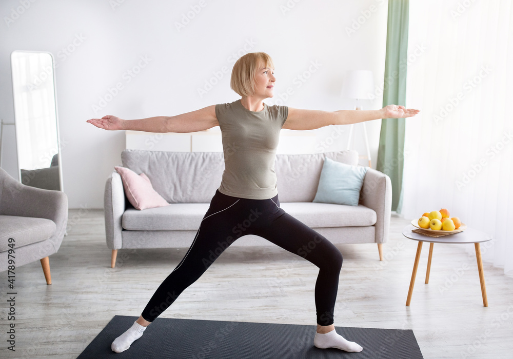 Strength yoga exercises. Fit mature lady standing in warrior asana, exercising on sports mat at home Strength yoga exercises. Fit mature lady standing in warrior asana, exercising on sports mat at home
