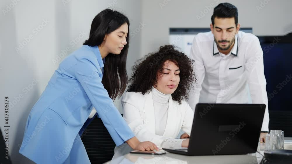 Group of confident multiracial colleagues discussing startup in office. Beautiful Caucasian and Asian women and Middle Eastern man talking and using online presentation on laptop. Teamwork concept. Group of confident multiracial colleagues discussing startup in office. Beautiful Caucasian and Asian women and Middle Eastern man talking and using online presentation on laptop. Teamwork concept.