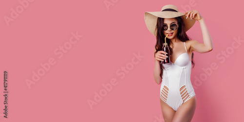 Fototapeta Tanned young woman in straw hat, white swimsuit hold a cocktail on pink background, isolate
