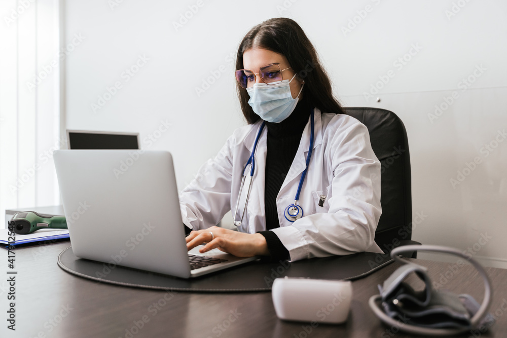 Young doctor working on laptop in office analyzes medical records of his patients infected whit Coronavirus Covid-19 wearing protective face mask - Professional sitting at desk looks at the screen Young doctor working on laptop in office analyzes medical records of his patients infected whit Coronavirus Covid-19 wearing protective face mask - Professional sitting at desk looks at the screen