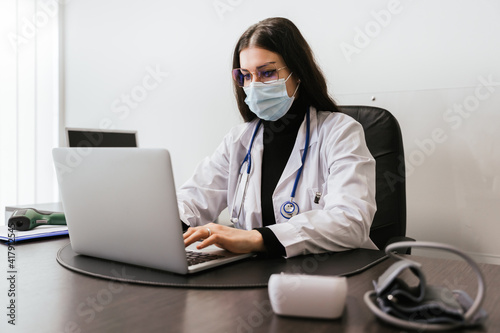 Young doctor working on laptop in office analyzes medical records of his patients infected whit Coronavirus Covid-19 wearing protective face mask - Professional sitting at desk looks at the screen