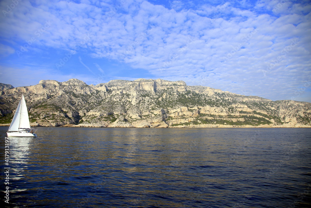 Overhanging rocky coast with scattered clouds in the blue sky and a sailboat, Parc National des Calanques, Marseille, France