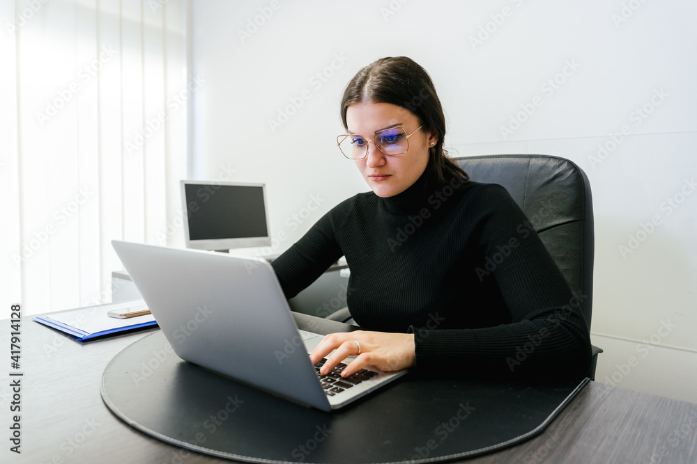 Successful woman sitting in office looks at work emails on her laptop and replies by typing on computer - Portrait of a serious busy secretary Successful woman sitting in office looks at work emails on her laptop and replies by typing on computer - Portrait of a serious busy secretary
