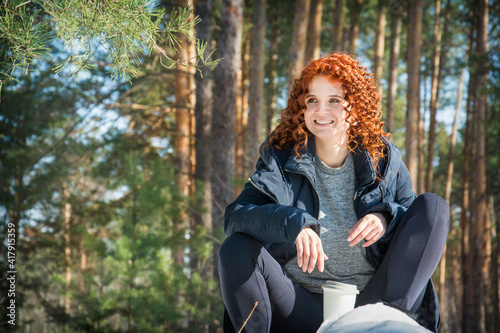 Beautiful redhead girl has a rest on the wooden log in winter