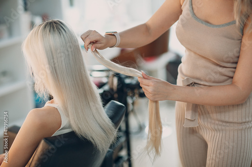 Hairdresser female making hair extensions to young woman with blonde hair in beauty salon. Professional hair extension. photo