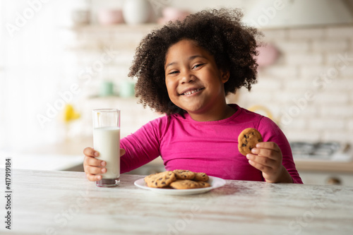 Tasty Snack. Cheerful Black Girl Eating Cookies And Drinking Milk In Kitchen